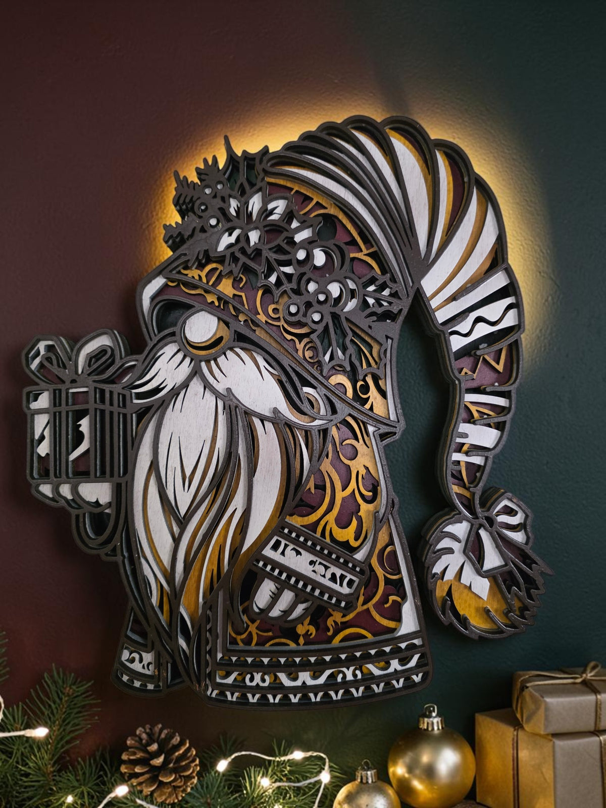 Layered wooden Christmas gnome wall art holding a wrapped gift, styled with evergreen branches, ornaments, pinecones, and warm holiday lights