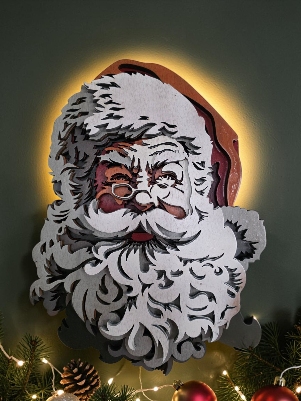 Layered wooden classic Santa Claus wall art styled with evergreen branches, ornaments, pinecones, and warm Christmas lights