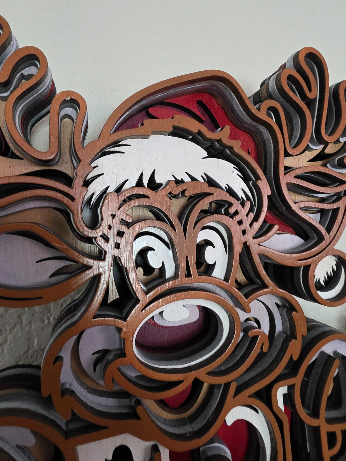 Close-up detail of smiling reindeer face, antlers, and Santa hat on layered wooden Christmas wall art